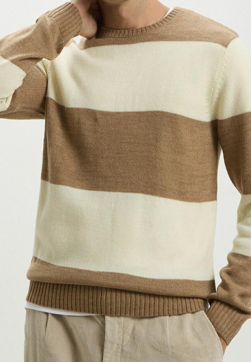 Homme portant un pull en tricot à rayures horizontales beige et crème avec un pantalon en velours côtelé beige, main reposant près du cou.