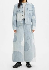 Chaqueta de mezclilla de color azul claro y pantalones de pierna ancha con grandes parches circulares blancos y en un azul más claro. Botas negras hasta el tobillo completan el conjunto.