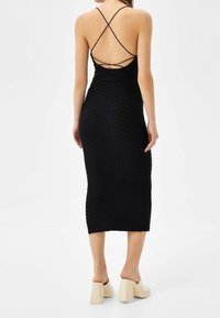 Schwarzes Bodycon-Kleid aus strukturiertem, geripptem Stoff. Verfügt über dünne, überkreuzte Träger und hat eine Länge bis zur Mitte der Wade. Kombiniert mit beigefarbenen Blockabsatzschuhen.