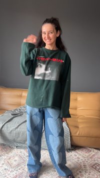 Zielony, oversize sweter z graficznym nadrukiem, w połączeniu z jasnoniebieskimi dżinsami z wzorem serc. W tle beżowa kanapa i kraciasta kołdra.