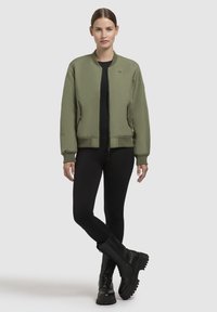 Olijfgroene bomberjack met een ronde halslijn en ritssluiting aan de voorkant, gecombineerd met zwarte legging en zwarte veterschoenen. Gladde textuur, minimalistisch ontwerp.