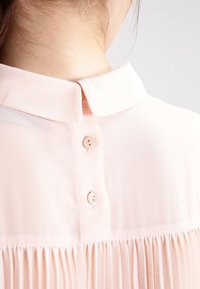 Blouse rose pâle avec un col doux, une fermeture à boutons au dos et une texture plissée le long de l'ourlet. Le tissu lisse rehausse son apparence.