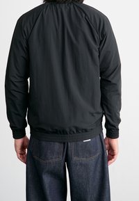 Svart lätt bomberjacka med ribbmuddar och elastisk midja, med en slät textur och avslappnad design. Bärs över mörka denimbyxor.