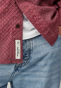 Chemise à manches longues à motifs rouges avec une étiquette indiquant "DURABLE GOODS", assortie à un jean en denim bleu clair. Le tissu a une finition texturée.