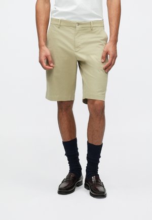 Shorts kaki cu croială dreaptă, închidere cu nasture și fermoar, buzunare laterale, purtați cu șosete din bumbac închis la culoare și mocasini maronii.