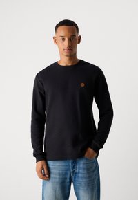Mandlig model iført en sort langærmet skjorte med et lille brun plet på brystet og blå jeans, stående foran en hvid baggrund.