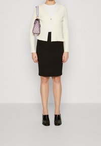 Cremfarbener gerippter Cardigan mit einem kleinen Knopf, weißes Oberteil, schwarzer Bleistiftrock und schwarze Slipper. Lila karierte Handtasche.