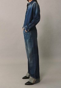 Camisa de denim y jeans de pierna ancha en azul oscuro. La camisa presenta un diseño de botones, mientras que los jeans tienen un lavado claro y dobladillos rematados con cremalleras.