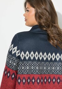 Cardigan lavorato a maglia con una parte superiore blu navy, una sezione inferiore rossa e intricati motivi geometrici bianchi in tutto. Morbido al tatto e vestibilità comoda.