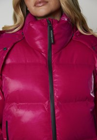 Fuksjowa kurtka puchowa z wysokim kołnierzem, z czarnym zamkiem błyskawicznym i guzikami oraz błyszczącą, pikowaną fakturą. Widoczna etykieta marki na zamku.