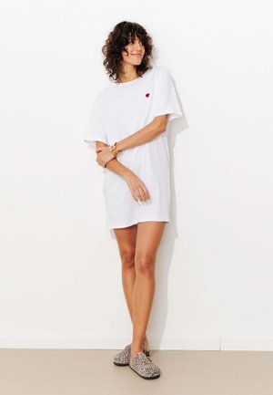 Femme aux cheveux bouclés portant une robe T-shirt blanche avec un petit cœur rouge, pantoufles imprimé léopard, debout contre un mur blanc.