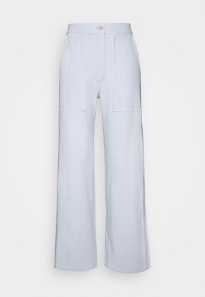 Punahiuksinen henkilö, jolla on beige lyhyt toppi, vaaleansiniset leveälahkeiset housut ja beiget korotetut slip-on-kengät, seisoo teksturoidun tuolin vieressä.