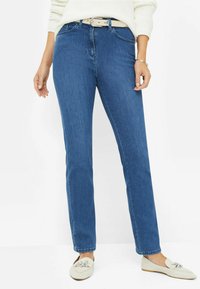Jean en denim bleu avec une coupe droite, taille haute, coutures discrètes et une ceinture beige. Assorti à des chaussures plates de couleur claire à enfiler.