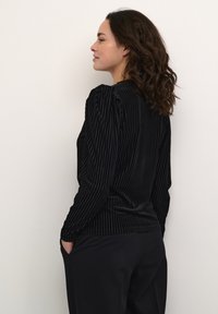 Juodas aksominis viršus su vertikaliomis juostelėmis, ilgomis rankovėmis ir laisvu siluetu. Pasižymi subtilia pūstuma peties siūlėje ir šoninėmis kišenėmis.
