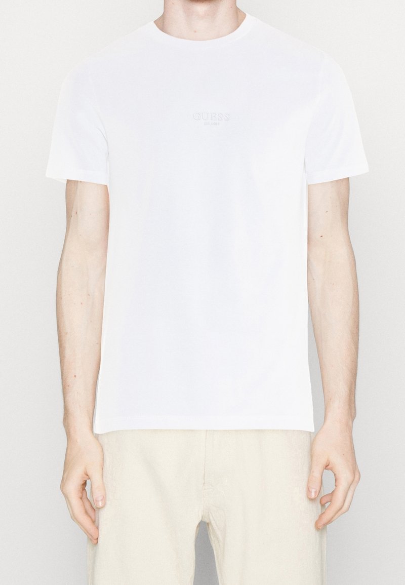 T-shirt bianco a maniche corte con logo discreto "GUESS EST. 1981" sul petto, indossato da una persona con pantaloni beige, in piedi davanti a uno sfondo neutro.