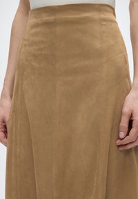 Κοντινό πλάνο ενός ατόμου που φοράει μια ψηλόμεση φούστα από ανοιχτό καφέ σουέτ με εμφανείς κάθετες ραφές και τα χέρια ακουμπισμένα στα πλάγια.