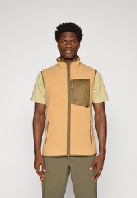 Tannenkleurige fleece vest met hoge kraag, voorzien van een groene ritszak en oranje ritslippen; gedragen over een lichtgroen T-shirt met korte mouwen.