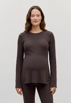 Mulher vestindo uma blusa justa canelada castanha de manga comprida e calças a condizer, em pé e a olhar para a frente contra um fundo claro e liso.
