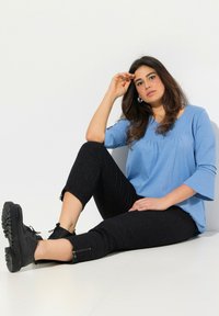 Hellblaues Langarmoberteil mit Falten, kombiniert mit schwarzen gemusterten Hosen, die über Reißverschlüsse an den Knöcheln verfügen. Schwarze Schnürschuhe.