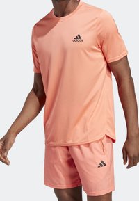 Camiseta deportiva de color melocotón con mangas cortas, cuello redondo y logo negro de Adidas; combinada con pantalones cortos a juego que cuentan con un bolsillo con cremallera.