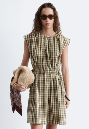 Femme portant des lunettes de soleil marron et une robe sans manches à carreaux verts tenant un chapeau beige tissé avec une écharpe marron à motifs.