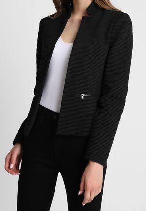 Femme portant un blazer noir texturé avec une poche zippée, par-dessus un top blanc et un pantalon noir, debout devant un fond uni.