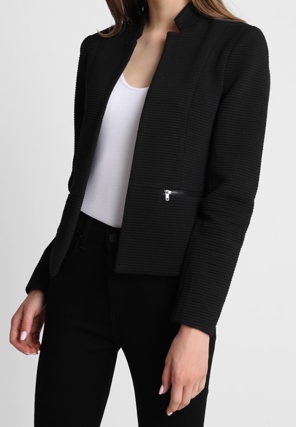 Femme portant un blazer noir texturé avec une poche zippée, par-dessus un top blanc et un pantalon noir, debout devant un fond uni.