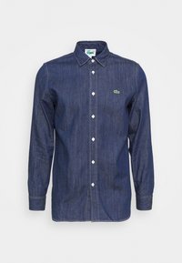 Mörkblå denimskjorta med lång ärm, knäppning framtill och vita knappar samt en liten grön broderad krokodil-logotyp på vänster bröst.