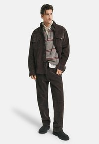 Homme portant une veste et un pantalon en daim brun foncé, un pull rayé gris, une chemise blanche et des chaussures noires, se tenant contre un fond blanc.