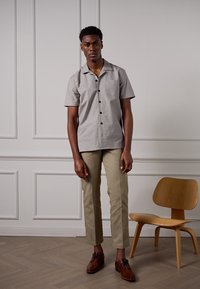 Chemise courte à manches courtes gris clair avec un devant à boutons, associée à un pantalon slim fit beige et des chaussures en cuir marron, le tout sur un fond uni.