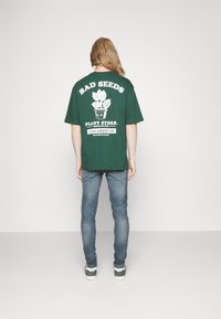 Camiseta de algodón verde de gran tamaño con texto gráfico blanco e ilustración de una planta en la parte de atrás, combinada con jeans de mezclilla azules y zapatillas.