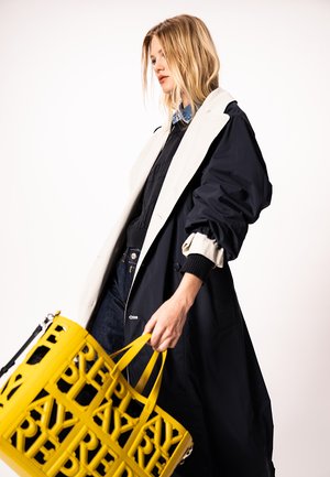 Szőke nő hosszú fekete és bézs kabátot visel, kezében egy nagy sárga táskát tart, amely kivágott betűmintával díszített.