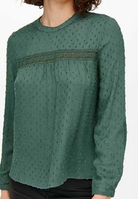 Grüne Bluse mit langen Ärmeln aus strukturiertem Stoff, mit einer Spitzenborte über der Brust und kleinen erhobenen Punkten im gesamten Design.