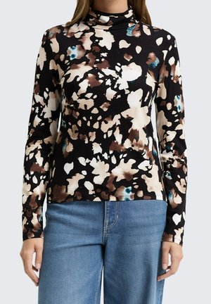 Kobieta ubrana w czarny top z golfem w brązowe, kremowe i niebieskie kwiatowe wzory z długimi rękawami, zestawiony z niebieskimi dżinsami.