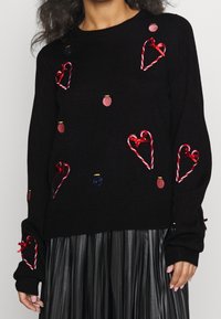 Svart stickad tröja med röda och guld broderade hjärtan och dekorationer, med inslag av pepparkaksgodis och röda rosetter på ärmarna.