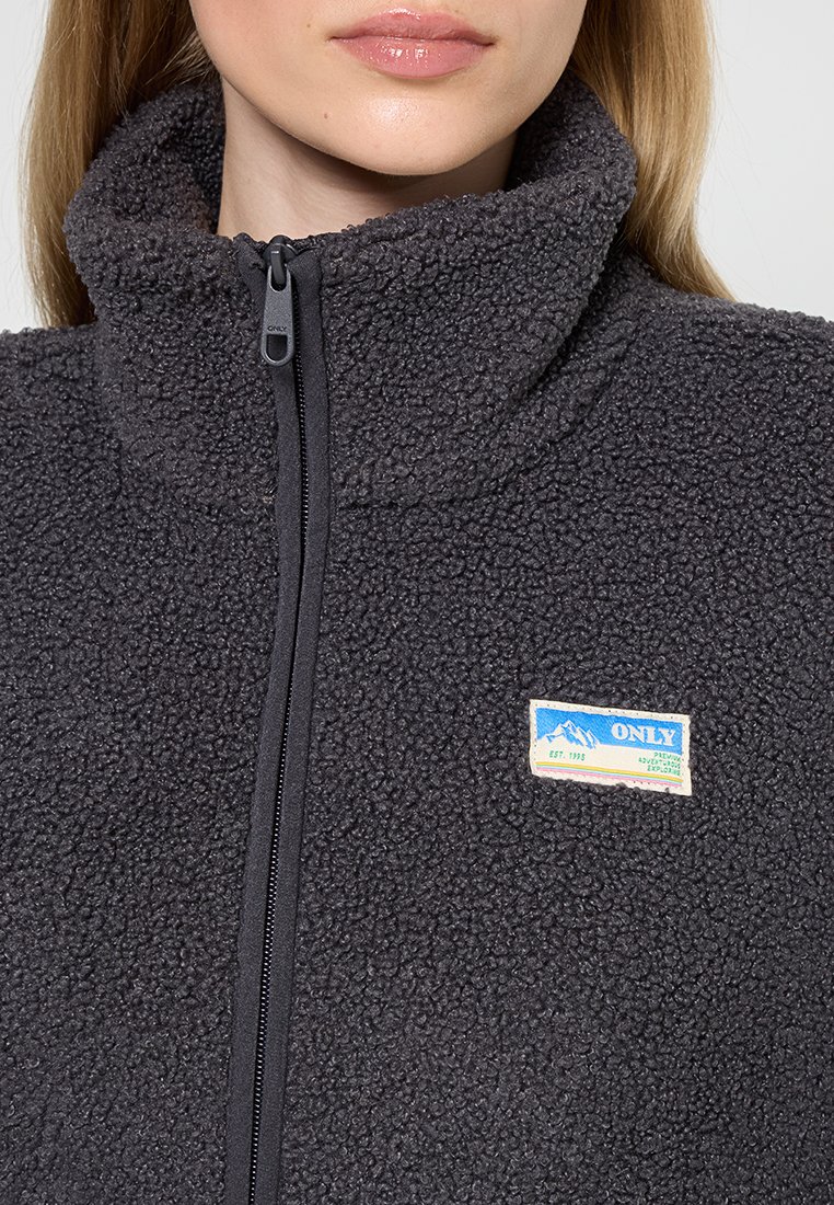 Dunkelgraue Fleecejacke mit hohem Kragen, vollständigem Reißverschluss und einem blauen und cremefarbenen Label auf der linken Brust. Weicher, strukturiert Stoff.