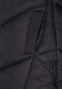 Prim-plan cu material de jachetă matlasată neagră, cu detaliu de petic dreptunghiular cusut și fermoar vizibil pe partea dreaptă.