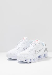 Ténis Nike Shox brancos com parte superior em malha, detalhes prateados e design distinto da sola com grandes pilares almofadados. Detalhe do logótipo em vermelho.