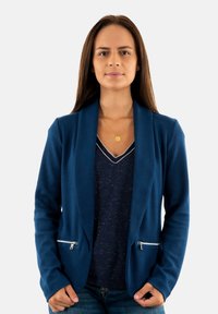 Blazer azul marino con cremallera y tejido texturizado, superpuesto a una blusa de pico azul oscuro. Diseño ajustado con mangas largas.