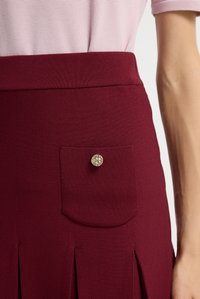 Burgundowa spódnica charakteryzuje się teksturowanym materiałem, tylną kieszenią z srebrnym guzikiem oraz plisami na dole dla dodatkowego wykończenia.
