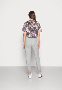 Mujer vistiendo un crop top floral colorido, pantalones de chándal grises claros y zapatillas blancas, de pie frente a un fondo claro y liso.