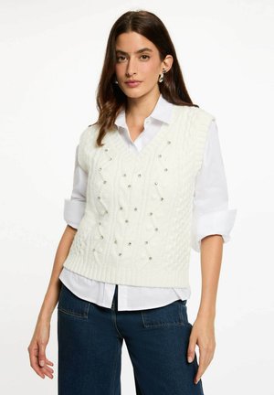 Gilet bianco lavorato a maglia con scollatura a V, un motivo testurizzato e perline decorative. Indossato sopra una camicia con colletto. Vestibilità over-size, maniche corte.