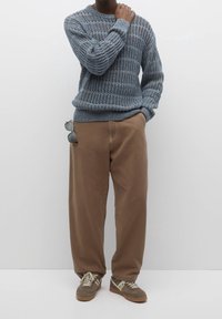 Person mit einem blauen Strickpullover und lockeren braunen Hosen, mit einer Sonnenbrille in der Tasche, stehend vor einem schlichten weißen Hintergrund.