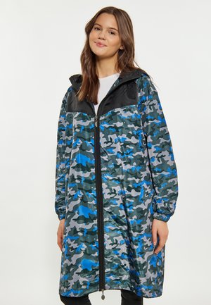 Junge Frau trägt eine lange blau-graue Camouflage-Zipjacke mit schwarzen Schulterpaneelen und elastischen Bündchen, steht vor weißem Hintergrund.