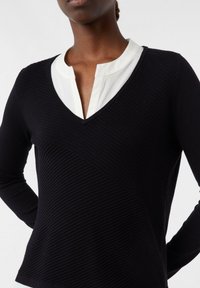 Frau trägt einen schwarzen gerippten V-Ausschnitt-Pullover über einem weißen Shirt mit rundem Kragen und Schlitz-Ausschnitt.