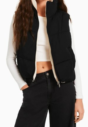 Vrouw draagt een zwart gewatteerd vest over een wit cropped top met lange mouwen en zwarte, hoge taille broek, met één hand in de zak.
