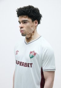 Camisola de futebol cinza claro com detalhes em bordo, apresentando um emblema, logóptico da marca e um acabamento riscado na gola e nas mangas.