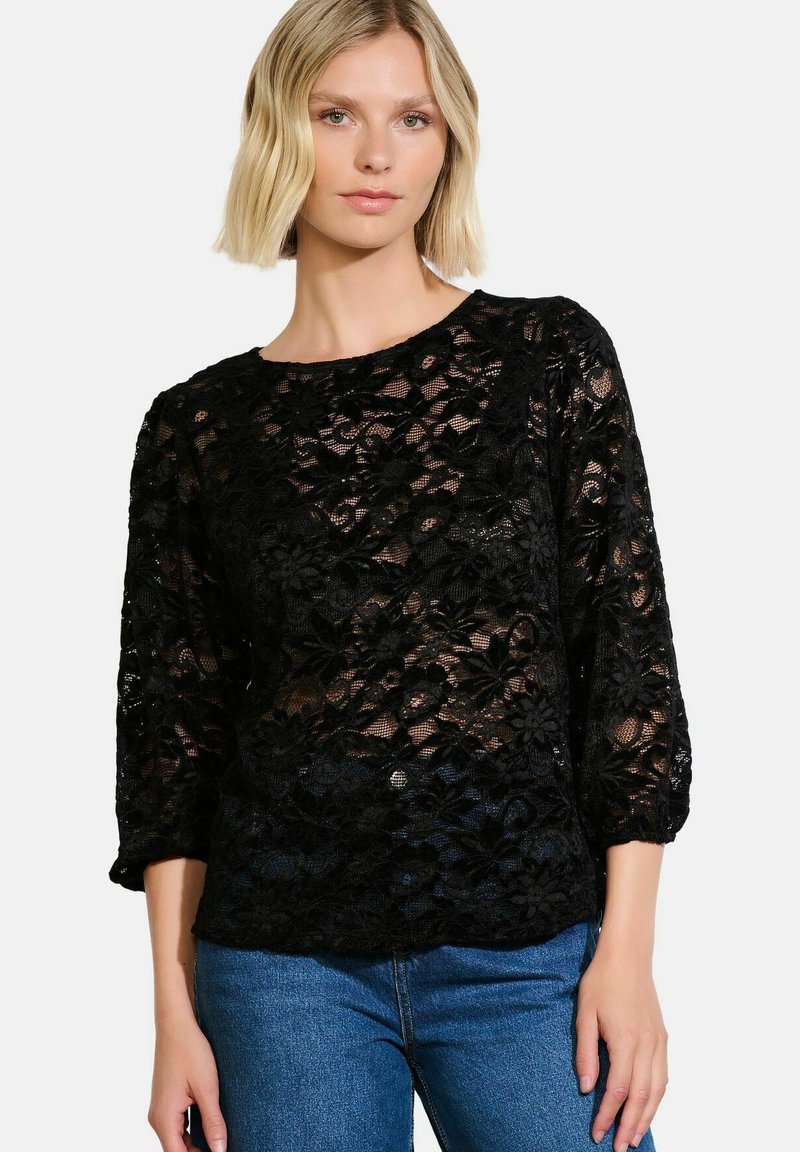 Juoda nėriniuota blusa su gėlių raštu, trijų ketvirčių rankovėmis, apvaliu iškirpimu ir permatomu audiniu, derinama su mėlynomis džinsinėmis kelnėmis.