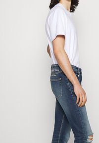 Jeans blu aderenti con un ginocchio strappato, abbinati a una semplice t-shirt bianca in cotone. I jeans presentano una vita con marchio e tasche posteriori.