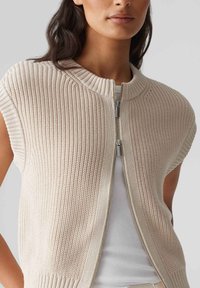 Stickad ärmlös cardigan i ljusbeige, med ribbad textur och silverfärgad dragkedja. Enklare rund halsringning och kortad design.